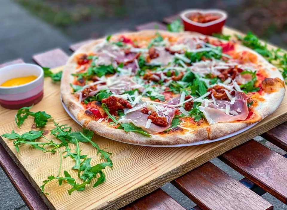 Pizza z rukolą i szynką na drewnianej desce, obok miseczka oliwy