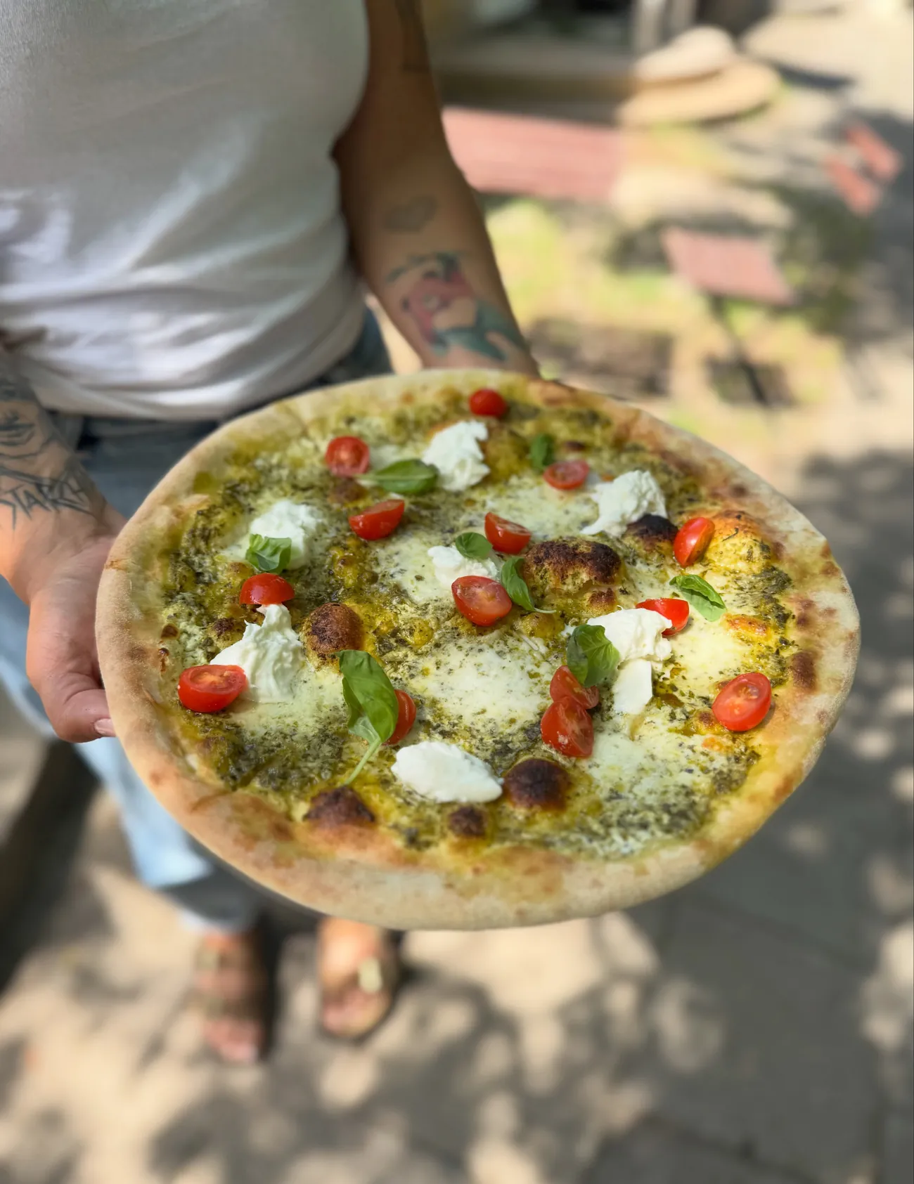 Pizza z pesto, bufalą i pomidorkami trzymana w dłoni, w tle ogródek