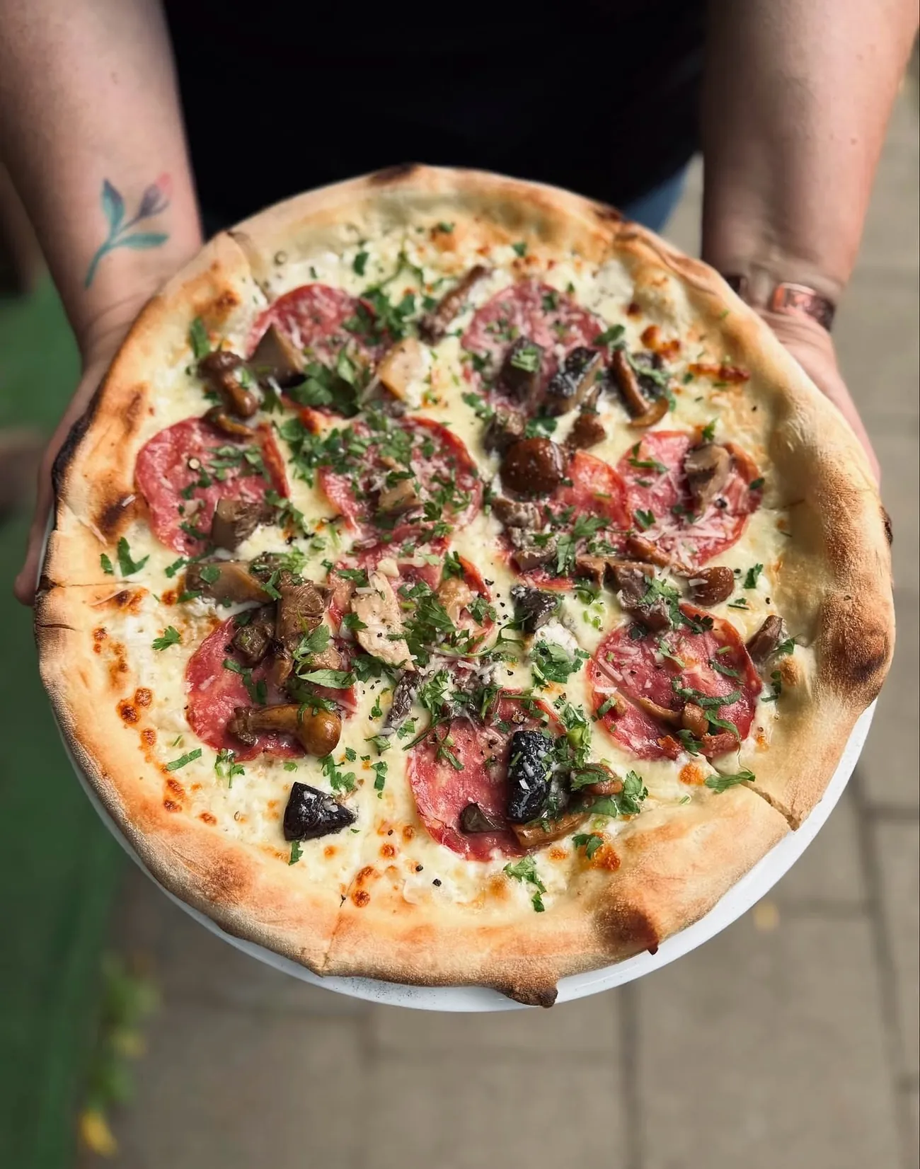 Pizza z salami, grzybami i natką pietruszki trzymana w wytatuowanych dłoniach